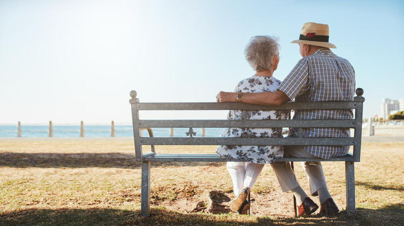 Rear view of loving senior couple relaxing at the seaside. Elderly man and woman sitting a bench outdoors.